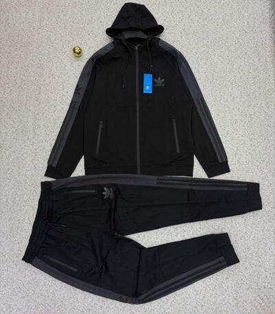 A stylish Adidas tracksuit laid out on a grey surface, featuring a black hoodie with grey accents and matching black pants with zipper pockets.