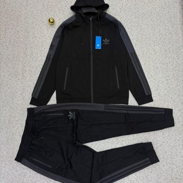 A stylish Adidas tracksuit laid out on a grey surface, featuring a black hoodie with grey accents and matching black pants with zipper pockets.