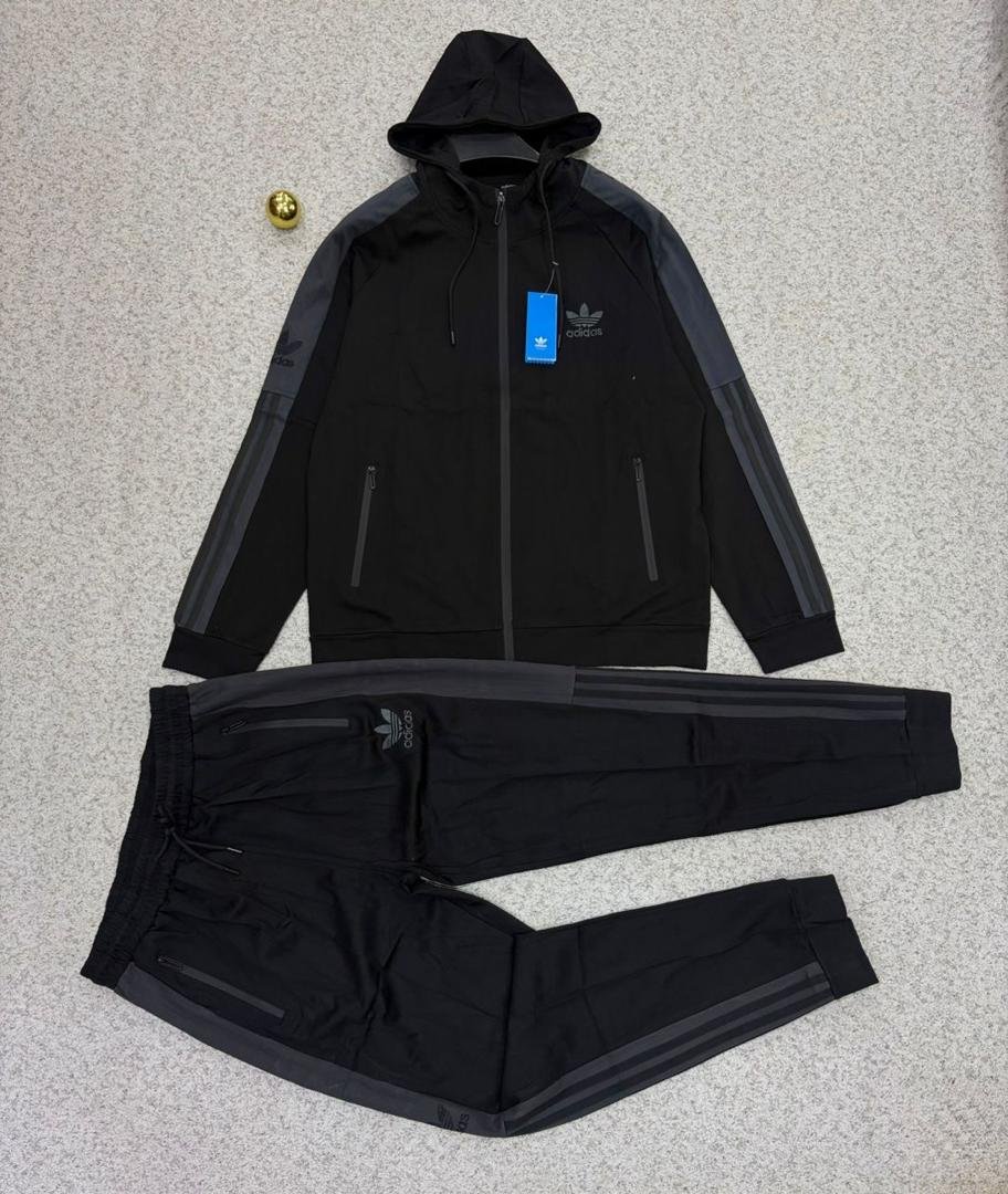 IMG-20260425-WA0068 A stylish Adidas tracksuit laid out on a grey surface, featuring a black hoodie with grey accents and matching black pants with zipper pockets.