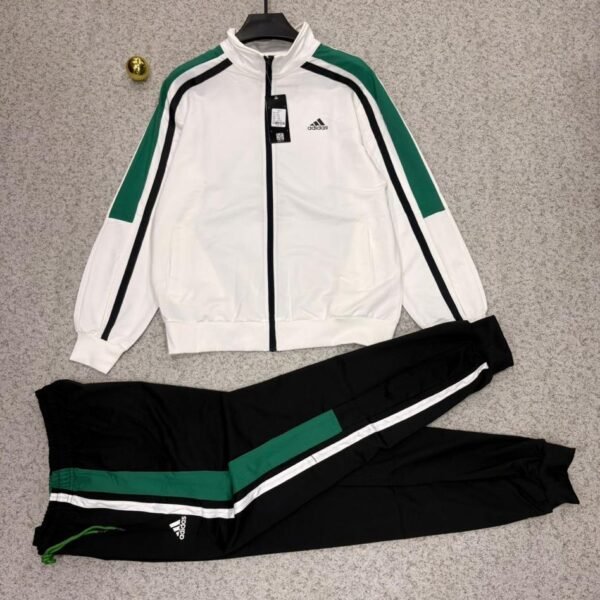 Adidas tracksuit laid out on a grey surface, featuring a white jacket with green and black stripes and matching black pants.
