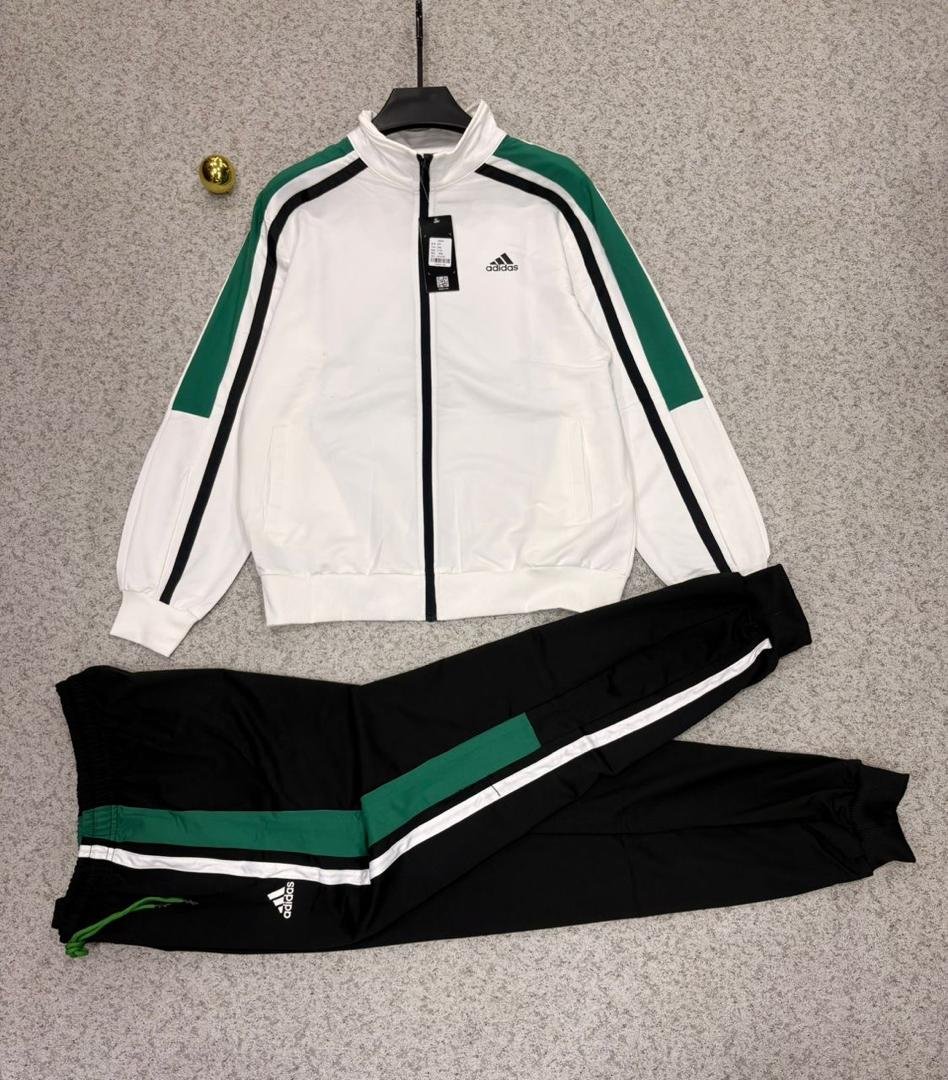 Adidas tracksuit laid out on a grey surface, featuring a white jacket with green and black stripes and matching black pants.