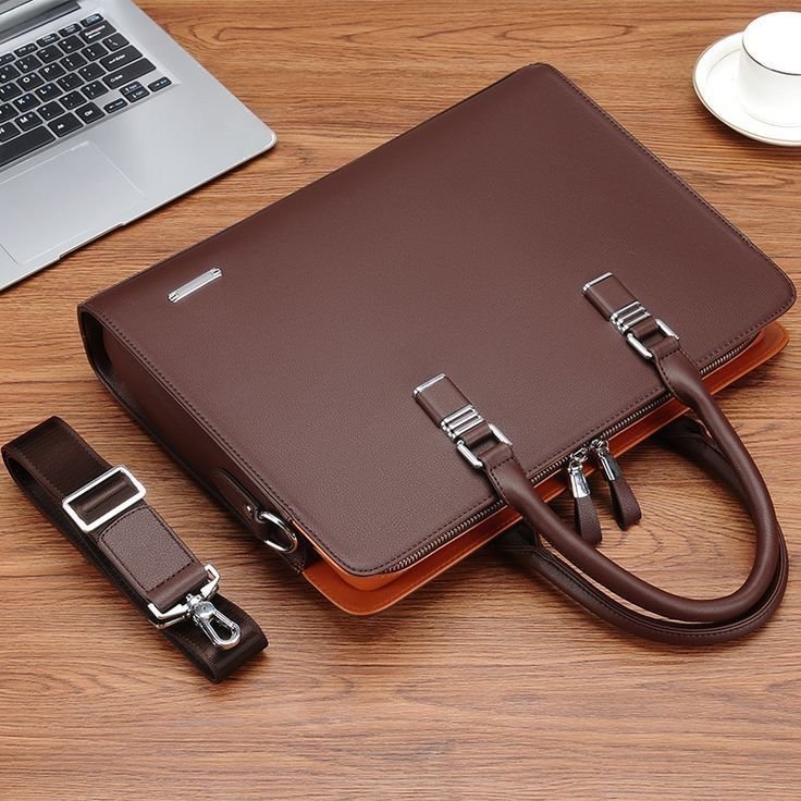 A stylish brown leather laptop briefcase with a detachable shoulder strap, placed on a wooden desk alongside a laptop and coffee cup.