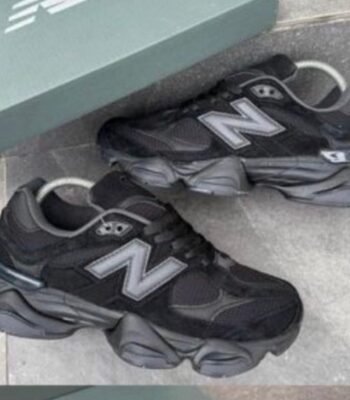 A pair of black and grey New Balance sneakers with white 'N' logo on a grey tile floor next to a green shoe box.