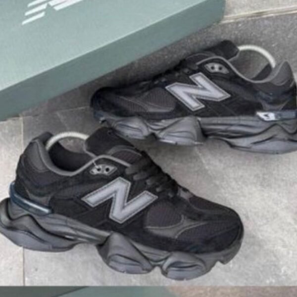 A pair of black and grey New Balance sneakers with white 'N' logo on a grey tile floor next to a green shoe box.