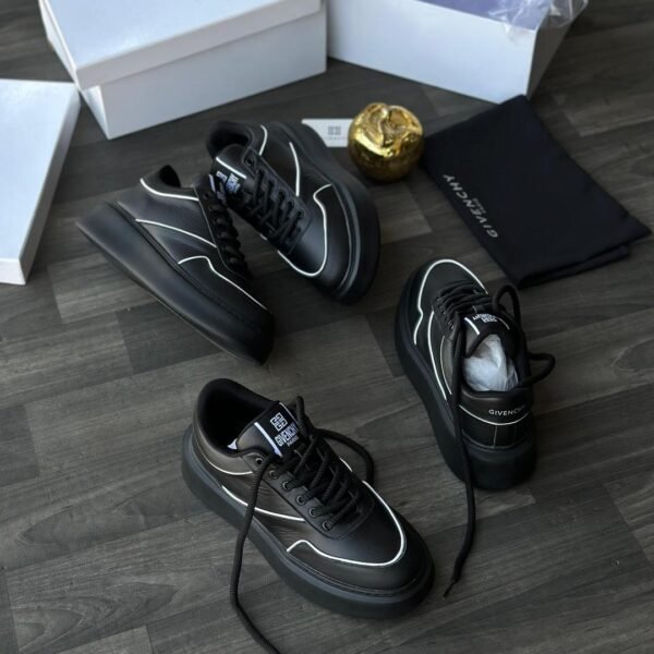 A pair of black leather Givenchy sneakers with white accents, displayed on a wooden floor with their box and packaging.