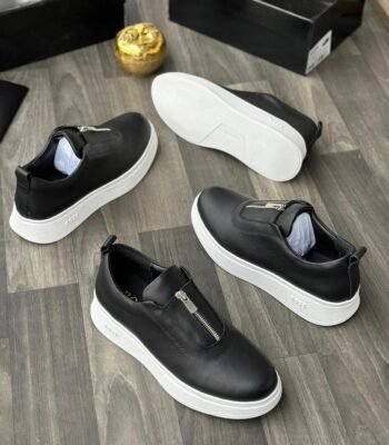 A pair of black leather sneakers with white soles and zip details, displayed on a wooden floor with a black shoe box and gold decorative item in the background.