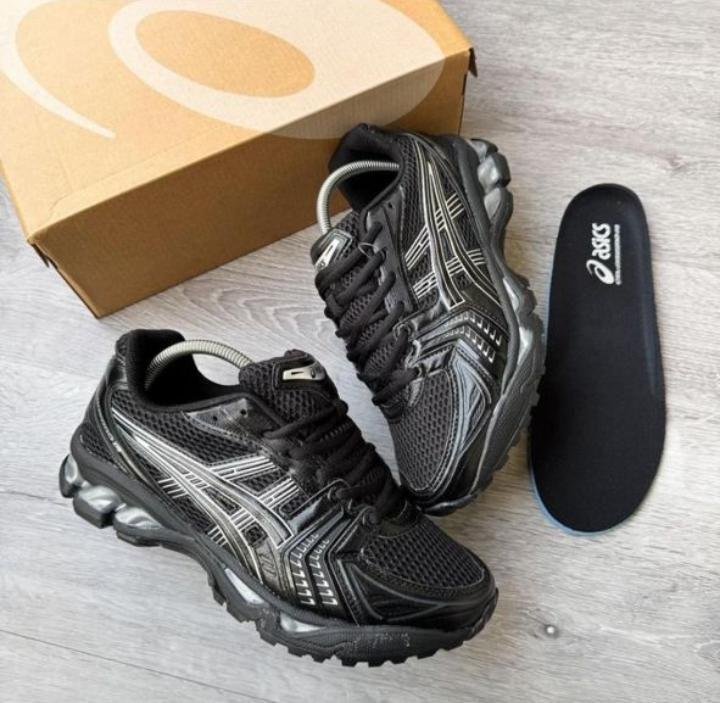IMG-20260425-WA0094 A pair of black and silver ASICS running shoes with a box and insoles on a light grey wooden floor, perfect for athletes seeking comfort and style.