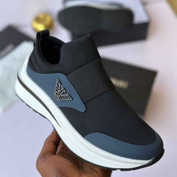 A close-up shot of a hand holding a black sneaker with a white sole and elasticated strap, showcasing the shoe's sleek design and luxurious details.