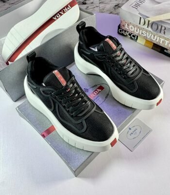 A pair of black Prada sneakers with red accents and white soles, placed on top of a Prada shoe box with luxury fashion books in the background.
