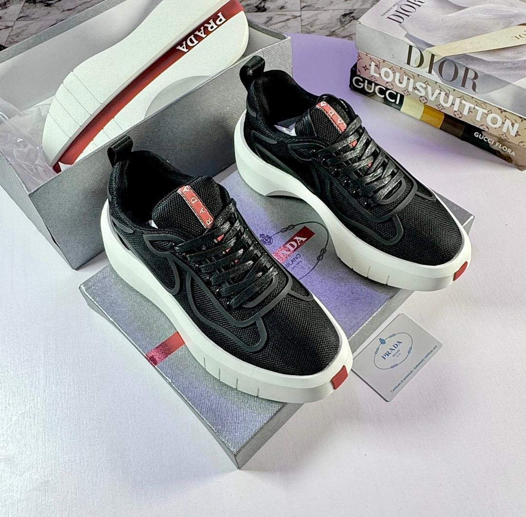 IMG-20260515-WA0012 A pair of black Prada sneakers with red accents and white soles, placed on top of a Prada shoe box with luxury fashion books in the background.