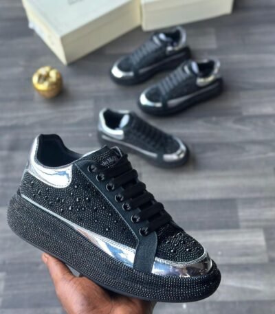 A close-up of a hand holding a black diamond-encrusted sneaker with silver accents, with two more pairs in the background on a grey wooden floor.