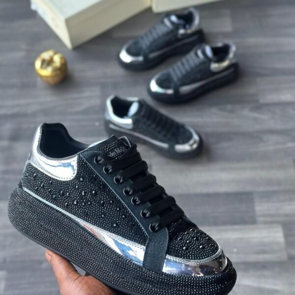 A close-up of a hand holding a black diamond-encrusted sneaker with silver accents, with two more pairs in the background on a grey wooden floor.