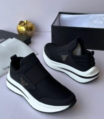 A pair of black and white Versace sneakers with gold accents, displayed on a white surface with a black box and gold accessories in the background.