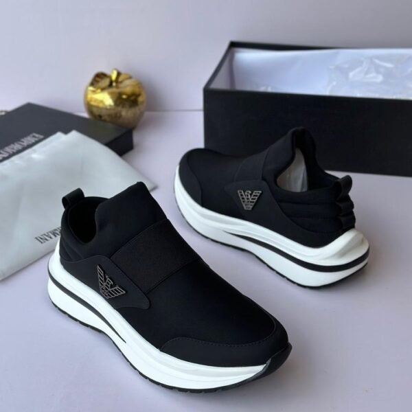 A pair of black and white Versace sneakers with gold accents, displayed on a white surface with a black box and gold accessories in the background.