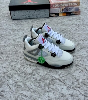 A pair of white and grey Air Jordan 4 sneakers with black accents, displayed on a marble surface with a shoe box and designer book covers in the background.