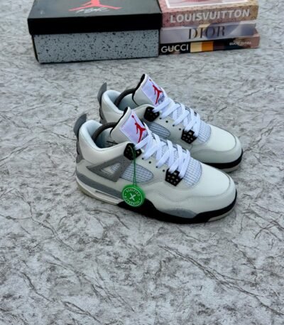 A pair of white and grey Air Jordan 4 sneakers with black accents, displayed on a marble surface with a shoe box and designer book covers in the background.