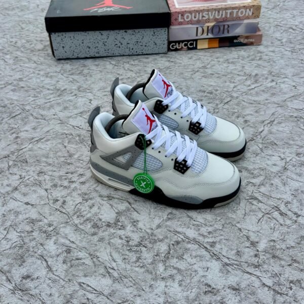 A pair of white and grey Air Jordan 4 sneakers with black accents, displayed on a marble surface with a shoe box and designer book covers in the background.