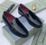 A pair of black crocodile pattern loafers with red interior, displayed on a shoe box with a luxurious background.
