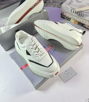 A pair of white Prada sport shoes with red accents and black detailing, placed on shoe boxes with a stack of luxury brand books in the background.