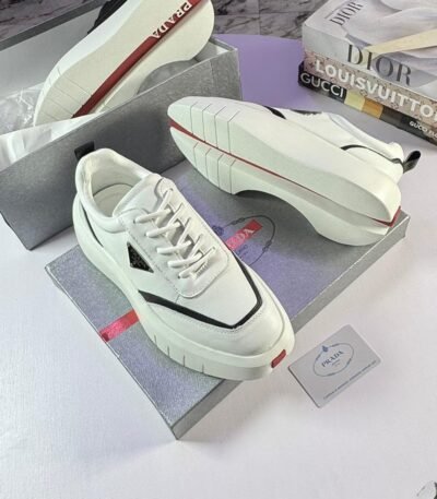 A pair of white Prada sport shoes with red accents and black detailing, placed on shoe boxes with a stack of luxury brand books in the background.