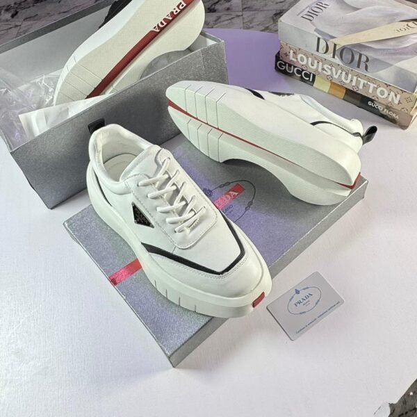 A pair of white Prada sport shoes with red accents and black detailing, placed on shoe boxes with a stack of luxury brand books in the background.