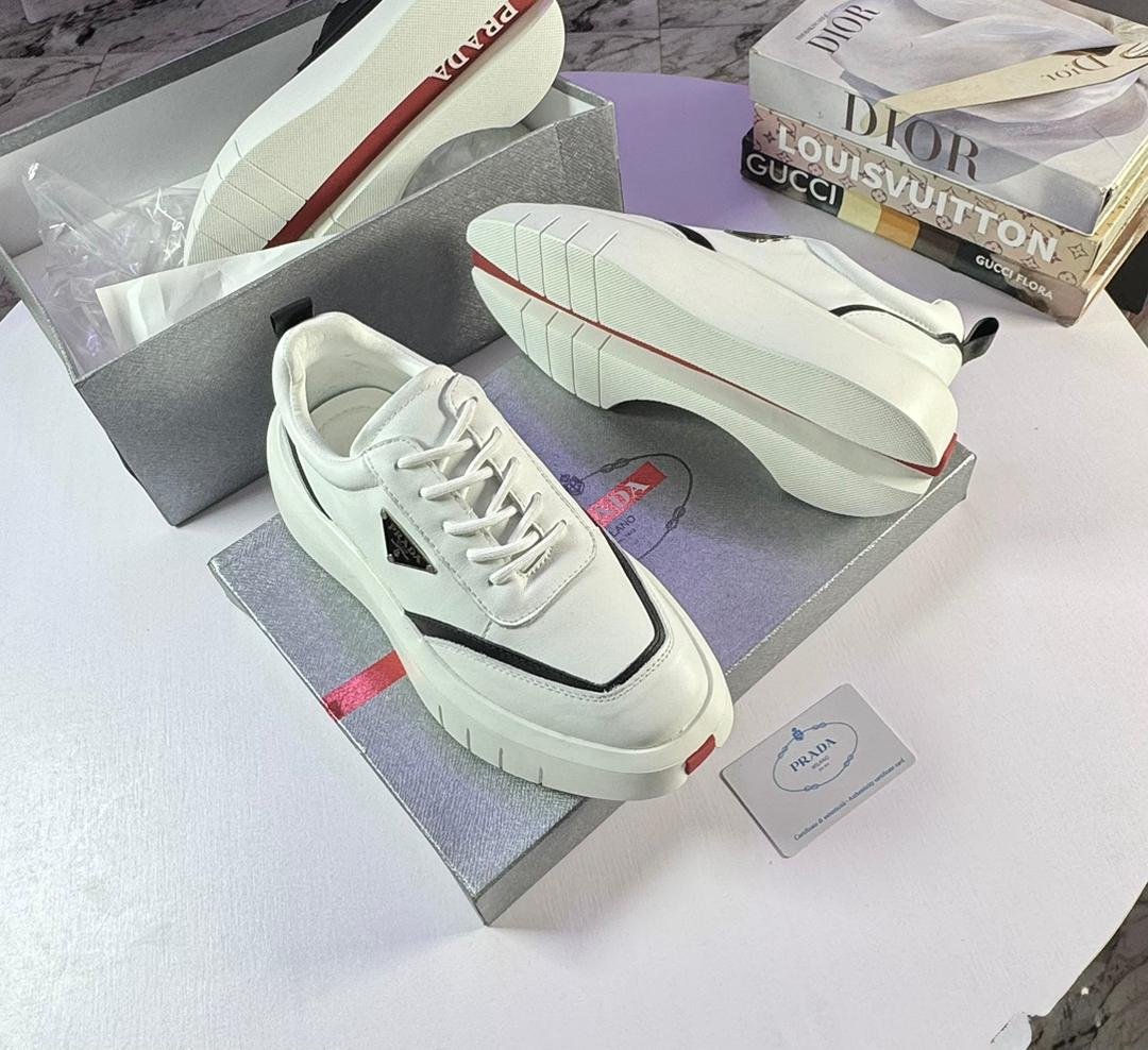 root A pair of white Prada sport shoes with red accents and black detailing, placed on shoe boxes with a stack of luxury brand books in the background.