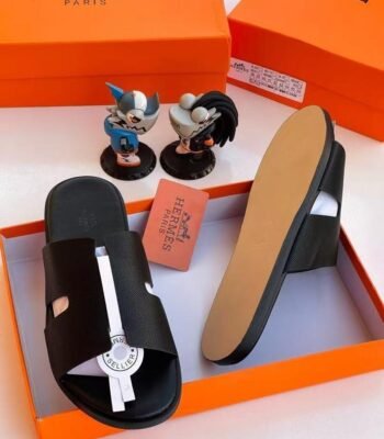 A pair of black leather slide sandals with a white accent strap, displayed in an orange Hermes Paris box with two figurines and a pink card.
