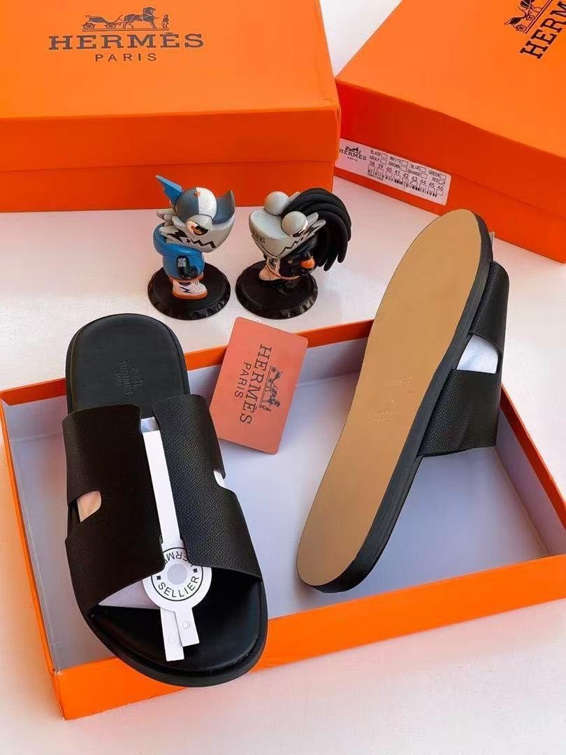 root A pair of black leather slide sandals with a white accent strap, displayed in an orange Hermes Paris box with two figurines and a pink card.