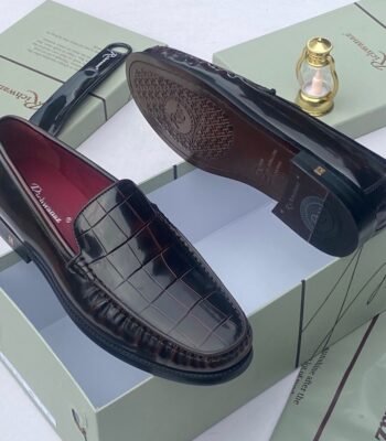 A pair of black crocodile pattern loafers with red interior, displayed on a shoe box with a luxurious background.