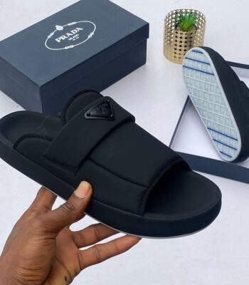 A hand holding a black Prada slide sandal with the shoe box and sole in the background, showcasing Modern Luxury and Urban Edge style.