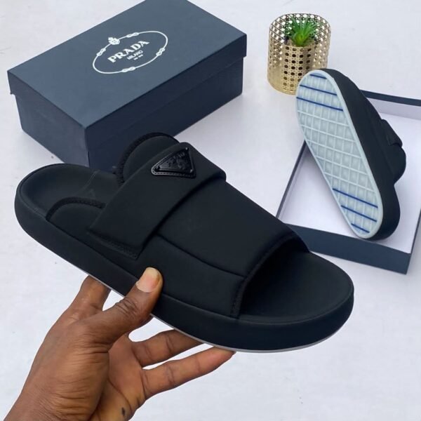 A hand holding a black Prada slide sandal with the shoe box and sole in the background, showcasing Modern Luxury and Urban Edge style.