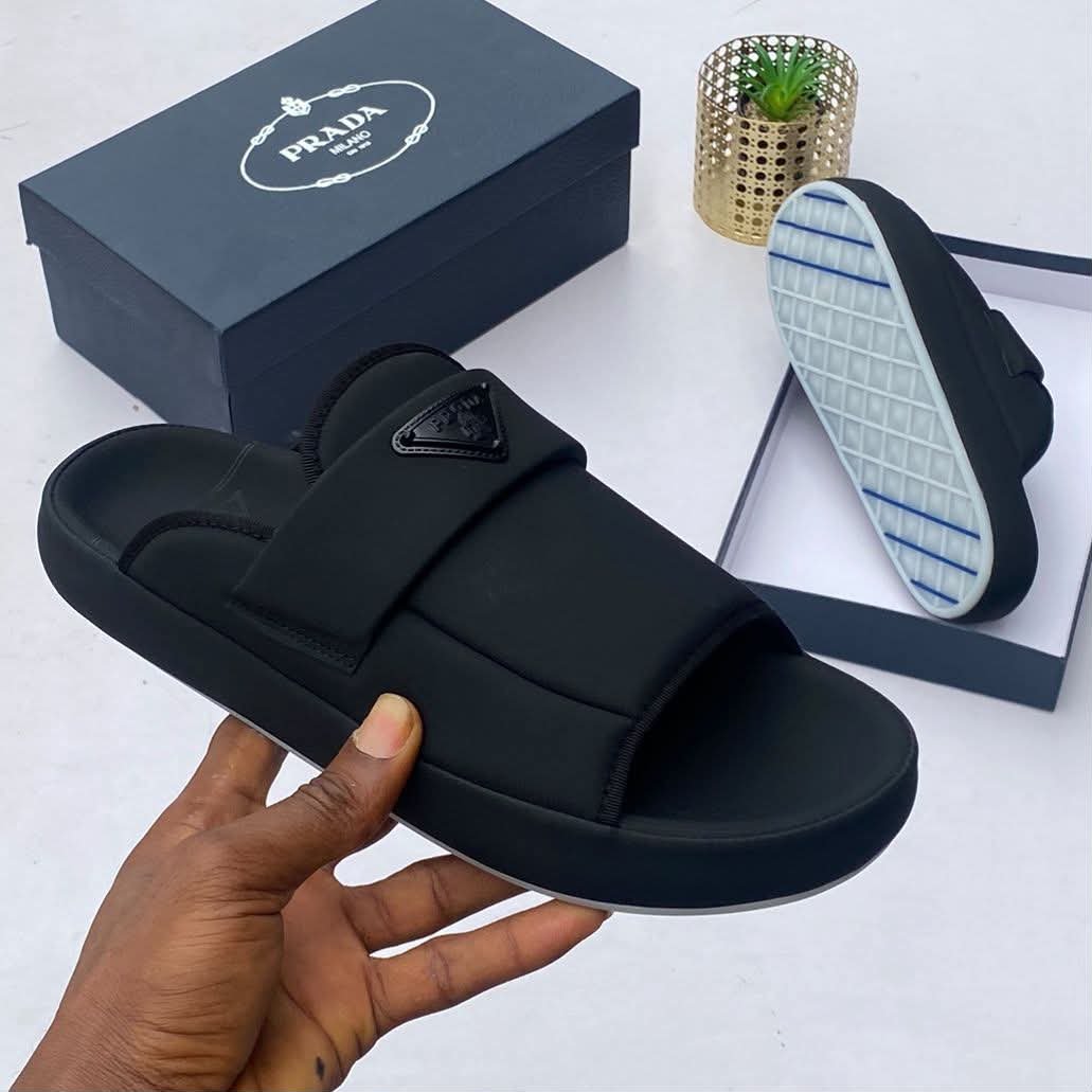 root A hand holding a black Prada slide sandal with the shoe box and sole in the background, showcasing Modern Luxury and Urban Edge style.