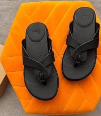 A pair of black two-strap flat sandals with embossed details, placed on an orange hexagonal cushion, exuding modern luxury and urban edge.