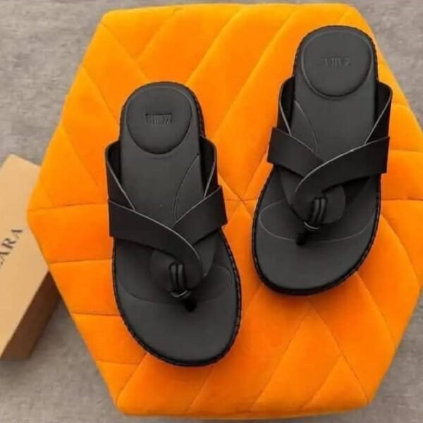A pair of black two-strap flat sandals with embossed details, placed on an orange hexagonal cushion, exuding modern luxury and urban edge.