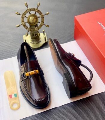 A pair of dark brown leather loafers with sleek buckle detail, presented on a white paper with a shoe horn and red shoe box in the background, embodying modern luxury and urban edge.