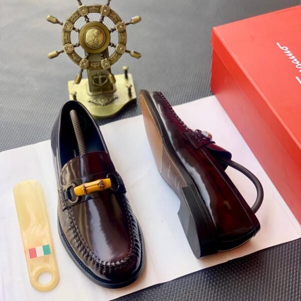 A pair of dark brown leather loafers with sleek buckle detail, presented on a white paper with a shoe horn and red shoe box in the background, embodying modern luxury and urban edge.