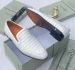 A pair of white leather loafers with perforated details, showcased on an open shoe box with elegant accessories, epitomizing luxury and sophistication.