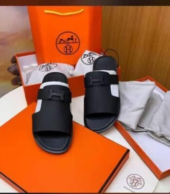 A pair of black Hermes rubber slides with double-buckle straps, displayed in an orange box and bag set, exuding luxury and sophistication.