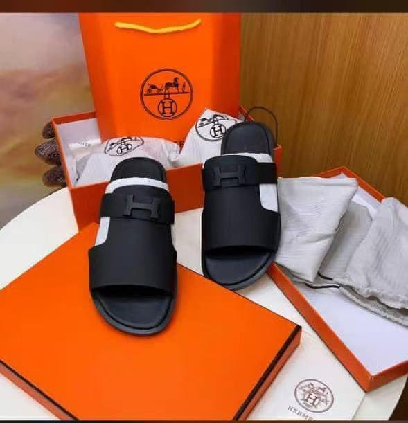 root A pair of black Hermes rubber slides with double-buckle straps, displayed in an orange box and bag set, exuding luxury and sophistication.
