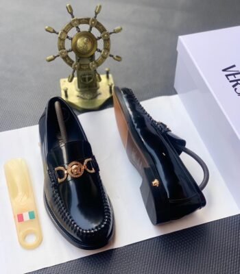 A pair of black leather loafers with a gold-tone Medusa buckle, presented on a white box with a shoe horn and a decorative ship's wheel in the background, embodying Modern Luxury and Urban Edge in Nigeria.