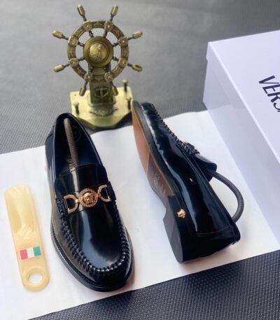 A pair of black leather loafers with a gold-tone Medusa buckle, presented on a white box with a shoe horn and a decorative ship's wheel in the background, embodying Modern Luxury and Urban Edge in Nigeria.