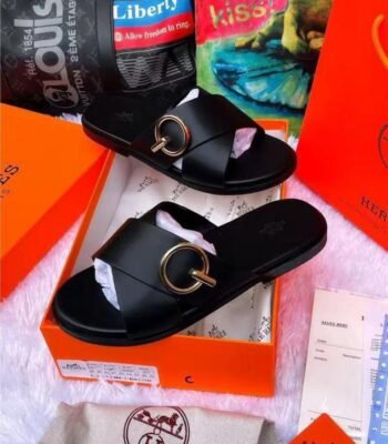 A pair of black leather slide sandals with a gold buckle, nestled in an orange box with luxury brand accessories, exuding modern luxury and urban sophistication.