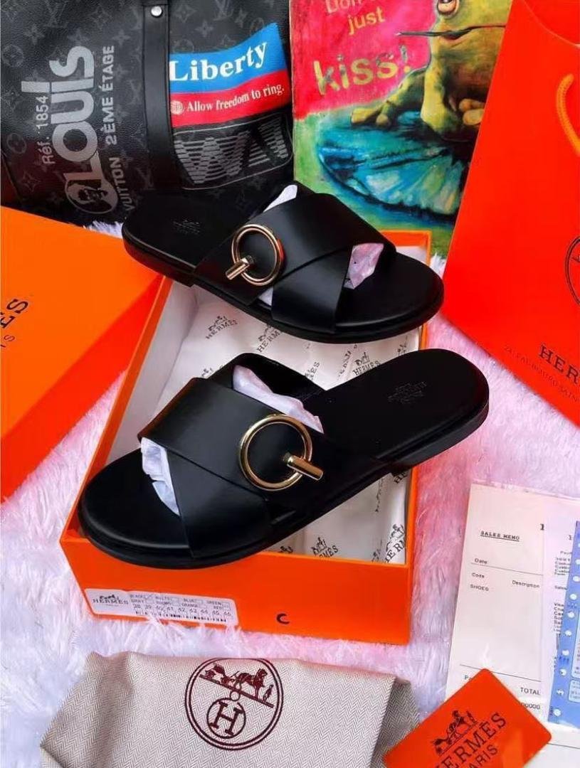 root A pair of black leather slide sandals with a gold buckle, nestled in an orange box with luxury brand accessories, exuding modern luxury and urban sophistication.