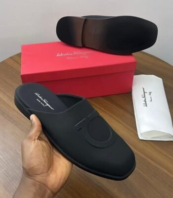 A pair of black leather mule loafers from Salvatore Ferragamo, displayed on a wooden table with the brand's signature red box and dust bag.