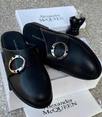 A pair of black leather mule shoes with a gold buckle from Alexander McQueen, displayed on a white box with the brand's logo, showcasing Modern Luxury and Urban Edge style.