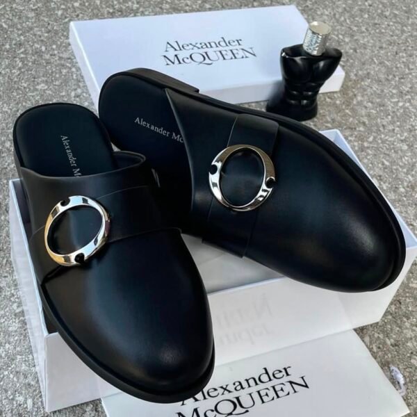 A pair of black leather mule shoes with a gold buckle from Alexander McQueen, displayed on a white box with the brand's logo, showcasing Modern Luxury and Urban Edge style.