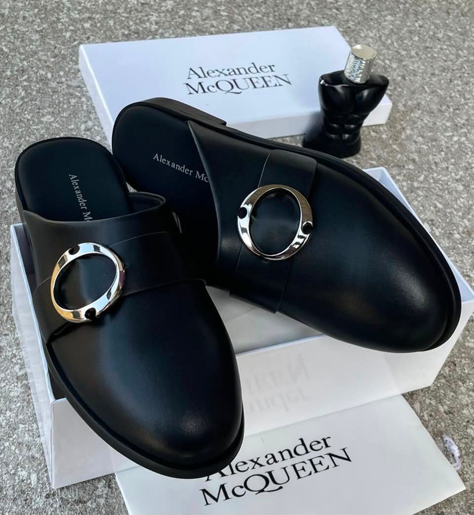 root A pair of black leather mule shoes with a gold buckle from Alexander McQueen, displayed on a white box with the brand's logo, showcasing Modern Luxury and Urban Edge style.