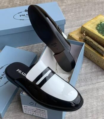 A pair of black patent leather half shoe with white accents by Prada, displayed in a blue shoe box on a light grey surface.