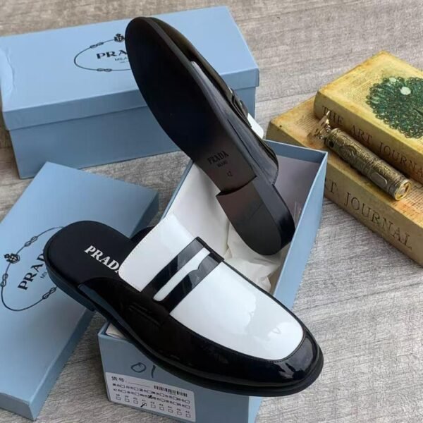 A pair of black patent leather half shoe with white accents by Prada, displayed in a blue shoe box on a light grey surface.