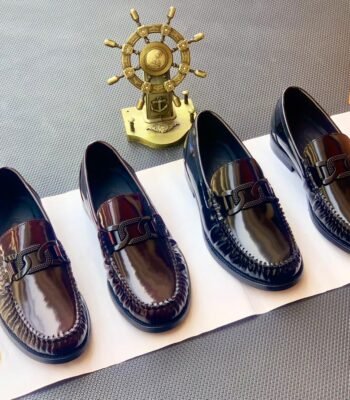 Four pairs of black leather loafers with sleek buckle detailing, showcasing modern luxury and urban edge, on a white paper with a golden ship's wheel in the background.